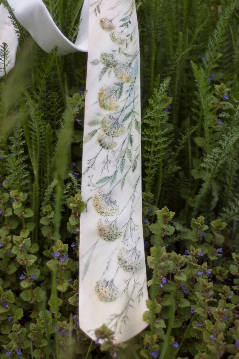 Puede incluir: Una corbata de seda blanca con un patr&oacute;n floral de hojas verdes y flores amarillas. La corbata est&aacute; sobre un lecho de plantas verdes.