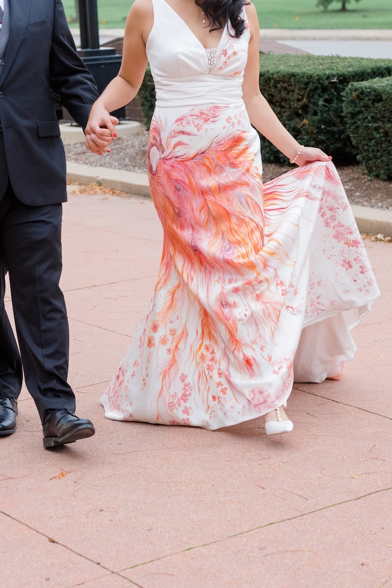 May include: A sleeveless white floor-length dress featuring a V-neckline and a hand-painted design of a large orange and red phoenix with flowing tail feathers and small pink floral accents extending across the skirt and bodice.