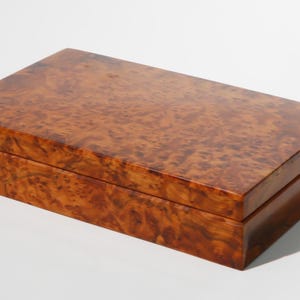 May include: A wooden box with a natural wood grain finish. The box has a hinged lid and is made of a light brown wood.