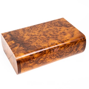 Puede incluir: Una caja rectangular de madera con un acabado pulido de veta de madera de raíz. La caja tiene bordes redondeados y un color marrón cálido con patrones intrincados. La tapa parece estar ligeramente entreabierta, revelando un vistazo del interior.