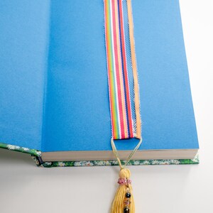 May include: A colorful ribbon bookmark is inserted into a blue-paged book. The bookmark features vertical stripes in various colors, including red, orange, yellow, green, and blue. A yellow tassel with beads and a silver fish charm hangs from the bookmark.