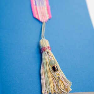 May include: A handmade bookmark with a colorful tassel and beaded accents. The bookmark features a pink and purple ribbon top, a tassel with pastel threads, and decorative beads, including a silver fish charm. The bookmark rests on a blue book.