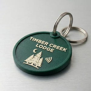 May include: A green circular keychain tag with the text "TIMBER CREEK LODGE" in white. The tag features a graphic of two pine trees and a crescent moon. Two silver key rings are attached to the tag.