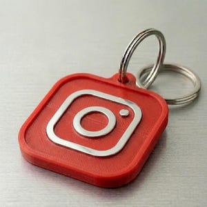 May include: A red Instagram logo keychain with a silver outline and a silver key ring. The keychain is square with rounded corners. The Instagram logo is in the center of the keychain.