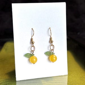 May include: A pair of gold-colored dangle earrings. Each earring features a yellow, round bead, a small green leaf, and gold-colored metal accents. The earrings are displayed on a white card.