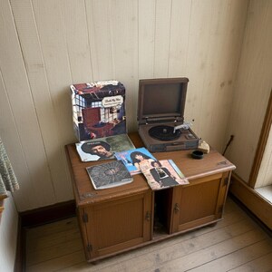 May include: A vintage wooden cabinet with a record player and several vinyl records. The record player is open, with a black record on the turntable. Album covers with images of musicians are visible. A box with a record player image is also present.