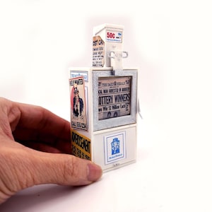 Può includere: Un piccolo distributore automatico di giornali bianco e argento con varie pubblicità. La macchina ha una fessura per 50 centesimi e mostra i titoli sui vincitori della lotteria. Una mano tiene la macchina.
