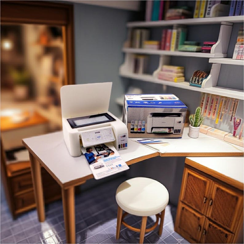 May include: Miniature white printer with a printed document emerging from the output tray. A box with the printer's image and ink cartridges sits nearby. The scene includes a small desk, stool, and a bookshelf, creating a dollhouse office setting.