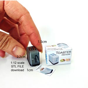 Peut inclure: Un grille-pain miniature argenté avec une base noire, mesurant 2,2 cm sur 1,5 cm. Le grille-pain est sur une surface blanche avec une petite boîte et une image imprimée du grille-pain. Le texte sur la boîte indique "TOASTER".