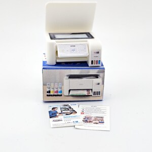 May include: A white Epson printer with an open top, sitting on a blue and white box. The printer has a small screen and colored ink cartridges. Two sheets of paper are in front of the printer.