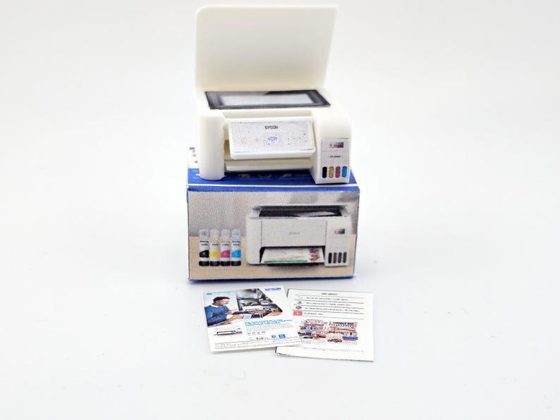 May include: A white Epson printer with an open top, sitting on a blue and white box. The printer has a small screen and colored ink cartridges. Two sheets of paper are in front of the printer.