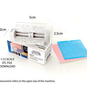 Może przedstawiać: Biały przecinacz do papieru z fakturowaną powierzchnią. Przecinacz ma 5 cm długości, 2,5 cm szerokości i 2 cm wysokości. Przecinacz znajduje się na pudełku z tekstem "Easycut Paper Cutter" i "1:12 Scale STL File Download".