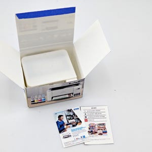 May include: An open white cardboard box with a blue-taped top, containing a white object. The box features a printed image of a printer and ink cartridges. Two leaflets with images and text are next to the box. The background is white.