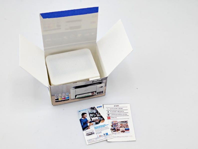 May include: An open white cardboard box with a blue-taped top, containing a white object. The box features a printed image of a printer and ink cartridges. Two leaflets with images and text are next to the box. The background is white.