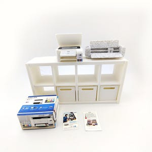 May include: A miniature white printer and a gray and white patterned cutting machine sit on a white shelf unit with eight cubbies. A printer box and two pamphlets are in front of the shelf.