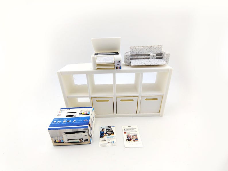 May include: A miniature white printer and a gray and white patterned cutting machine sit on a white shelf unit with eight cubbies. A printer box and two pamphlets are in front of the shelf.