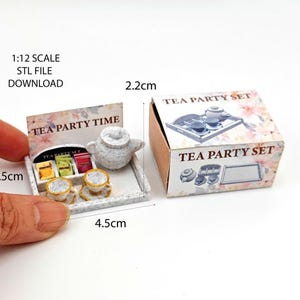 Puede incluir: Un juego de té en miniatura para una fiesta con una tetera, tazas y bolsitas de té en una bandeja. El juego mide 4,5 cm de ancho y 3,5 cm de alto. La caja mide 2,2 cm de alto y tiene el texto "Tea Party Set" en ella.