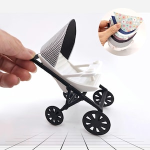 Puede incluir: Un cochecito blanco y negro en miniatura con marco y ruedas negros. El cochecito tiene una capota estampada y se muestra con una mano que sostiene varios recortes de papel estampados.