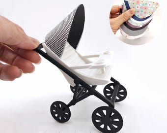 May include: A miniature white and black stroller with a black frame and wheels. The stroller has a patterned canopy and is shown with a hand holding several patterned paper cutouts.