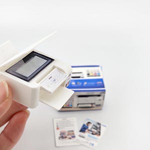 May include: A miniature white printer with a black screen and a small paper tray. The printer is held in a hand, with a box and two small brochures in the background. The printer is a model, likely for a dollhouse or miniature scene.