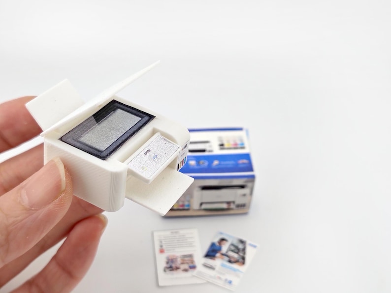 May include: A miniature white printer with a black screen and a small paper tray. The printer is held in a hand, with a box and two small brochures in the background. The printer is a model, likely for a dollhouse or miniature scene.
