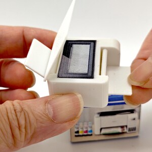 May include: A miniature white printer with a black screen and a hinged top. The printer is being held open by a person's fingers, revealing the interior. The printer is sitting on top of a box with a picture of a printer.