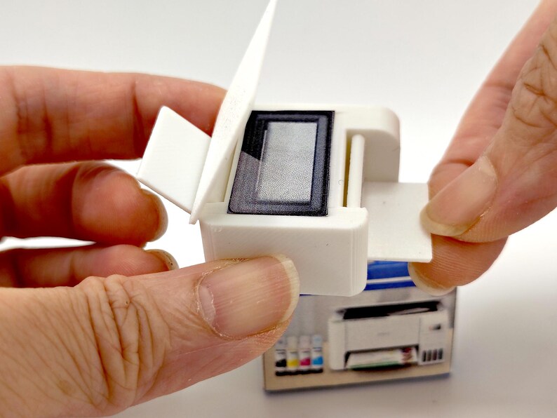 May include: A miniature white printer with a black screen and a hinged top. The printer is being held open by a person's fingers, revealing the interior. The printer is sitting on top of a box with a picture of a printer.