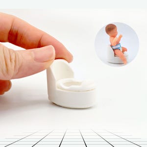 May include: A miniature white potty chair with a rounded back and seat. A small doll of a baby wearing a diaper is sitting on a similar potty chair in the background. The image is set against a white background.