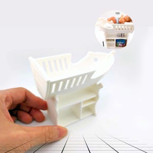 May include: A white miniature doll's crib with a boat-like design, featuring a slatted headboard and footboard. The crib has a storage shelf underneath. A hand is holding the crib. A smaller image shows a doll and accessories.