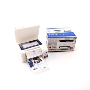 May include: A white Epson printer with a black display screen and a printed document emerging from the output tray. The printer is next to its original packaging, which features a photo of the printer and ink cartridges.