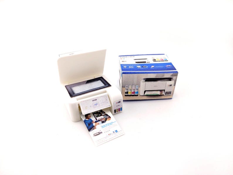 May include: A white Epson printer with a black display screen and a printed document emerging from the output tray. The printer is next to its original packaging, which features a photo of the printer and ink cartridges.