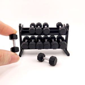 May include: Miniature black dumbbells with a silver handle, displayed on a black rack. One dumbbell is held by a hand. The weights are small, likely for a dollhouse or model. The rack has two tiers for storage.