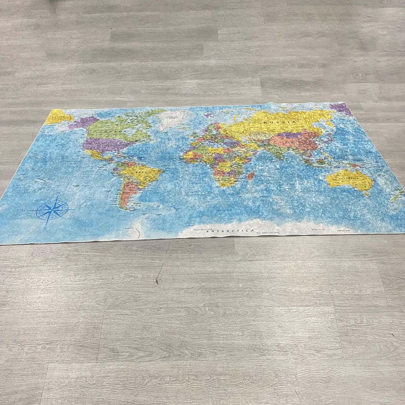 May include: A rectangular world map rug with a blue background and colorful continents. The map includes a compass rose in the lower left corner and the word "ANTARCTICA" at the bottom. The rug is lying on a light gray wooden floor.