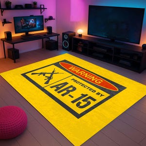 Puede incluir: Una alfombra rectangular amarilla con borde negro y el texto "WARNING PROTECTED BY AR-15". La alfombra presenta un letrero de advertencia rojo y siluetas de dos rifles. La alfombra está en una habitación con una computadora y un televisor.