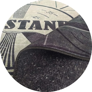 Pu&ograve; includere: Un tappeto circolare dal design vintage. Il tappeto presenta la parola "STAND" in lettere grandi e in grassetto, insieme a una grafica a forma di orologio e linee. Il bordo del tappeto &egrave; arricciato, rivelando un lato inferiore grigio scuro e strutturato.