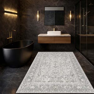 Può includere: Un tappeto rettangolare grigio chiaro con un motivo floreale ornato in un bagno moderno. Il tappeto è centrato sul pavimento grigio scuro, di fronte a un mobiletto in legno e una vasca da bagno nera. Il bagno ha infissi e illuminazione dorati.