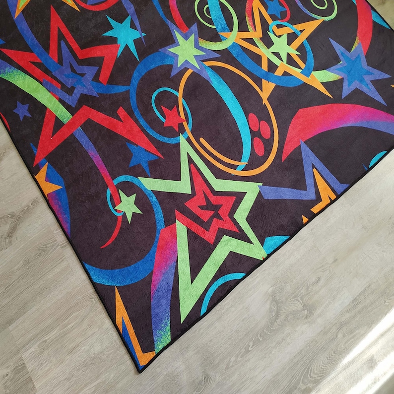 May include: A black rug with a colourful design featuring stars, swirls, and rainbow-like patterns. The rug has a dark background with bright red, green, blue, and orange accents.