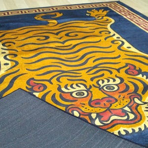 May include: A rectangular rug featuring a stylized tiger design. The tiger is primarily yellow and orange with black outlines, set against a dark blue background. The rug has a decorative border.