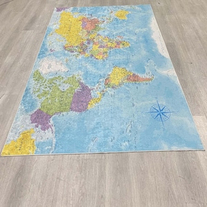 May include: A rectangular rug featuring a world map design. The map displays continents in various colors, including yellow, green, and purple, against a blue ocean background. A blue compass rose is in the lower right corner. The rug is placed on a light gray wooden floor.