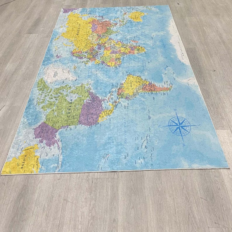 May include: A rectangular rug featuring a world map design. The map displays continents in various colors, including yellow, green, and purple, against a blue ocean background. A blue compass rose is in the lower right corner. The rug is placed on a light gray wooden floor.
