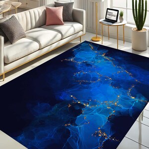 May include: A large rectangular rug with a dark blue and bright blue abstract design. The rug has gold flecks throughout the design, and is placed in a living room setting with a white sofa and pillows.