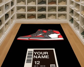 May include: A black rug featuring a red, gray, and white high-top sneaker design. The rug also includes a white label with the text "YOUR NAME" and size information: 12, UK 11, CM 30, BR 44, and EUR 46. The rug is in a shoe closet.