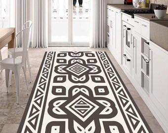 May include: A rectangular kitchen rug with a geometric pattern in shades of brown and white. The rug features a central design with diamond and floral shapes, bordered by triangles. It is placed in front of a white kitchen.