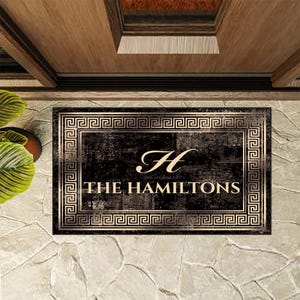 May include: A rectangular doormat with a black and distressed gold finish. The border features a repeating Greek key pattern. The center displays a large script letter H above the text THE HAMILTONS in a bold, serif, gold-colored font.