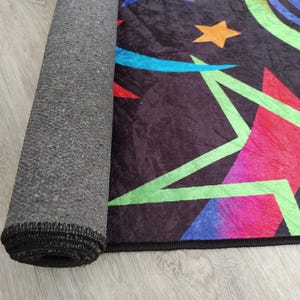 May include: A black rug with a colourful geometric pattern featuring stars, swirls, and stripes. The rug has a grey backing and black edging.