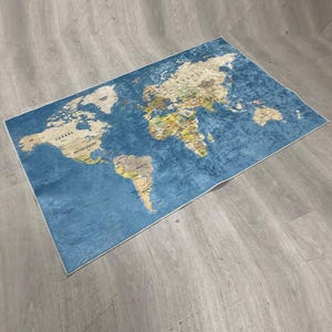 May include: A rectangular rug featuring a world map design. The rug has a blue background with a detailed map in shades of beige, yellow, and green. The map includes country names and borders, suitable for a living room or study.