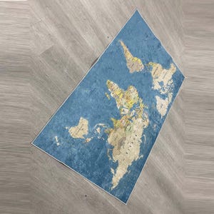 May include: A rectangular blue rug with a world map design. The map features continents in beige and countries in various colors. The rug is placed on a light gray wooden floor.
