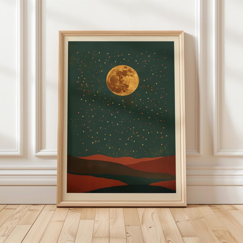 May include: A framed art print featuring a celestial landscape. The artwork depicts a large, golden moon and numerous stars against a dark teal sky. Below, there are layered red and brown hills. The print is encased in a light wood frame.