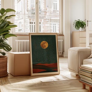 May include: A framed art print featuring a celestial landscape. The artwork depicts a large, textured, orange moon against a dark teal sky filled with small white stars. Below the sky are reddish-brown hills. The frame is a light wood color.