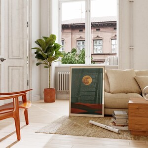 May include: A framed art print featuring a full moon over a landscape with a ladder. The artwork has a dark green background with a yellow moon and red and brown hills. The print is displayed in a bright, airy room with a wooden chair and a large potted plant.
